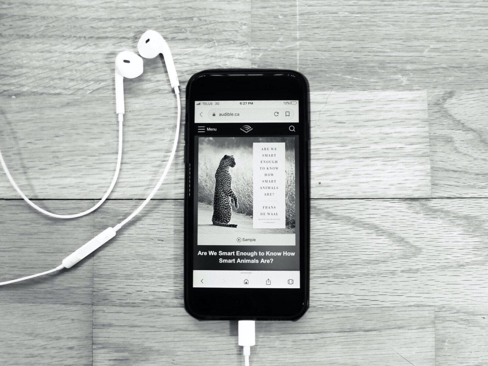 Black and white photo of a smartphone displaying the audiobook "Are We Smart Enough to Know How Smart Animals Are?" by Frans de Waal on the Audible app. Earbuds are also visible in the image.