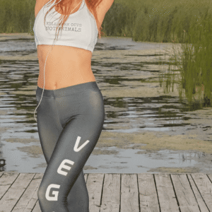 A woman wearing black leggings with the word "VEGAN" printed vertically on the side, stretching on a wooden dock overlooking a lake.