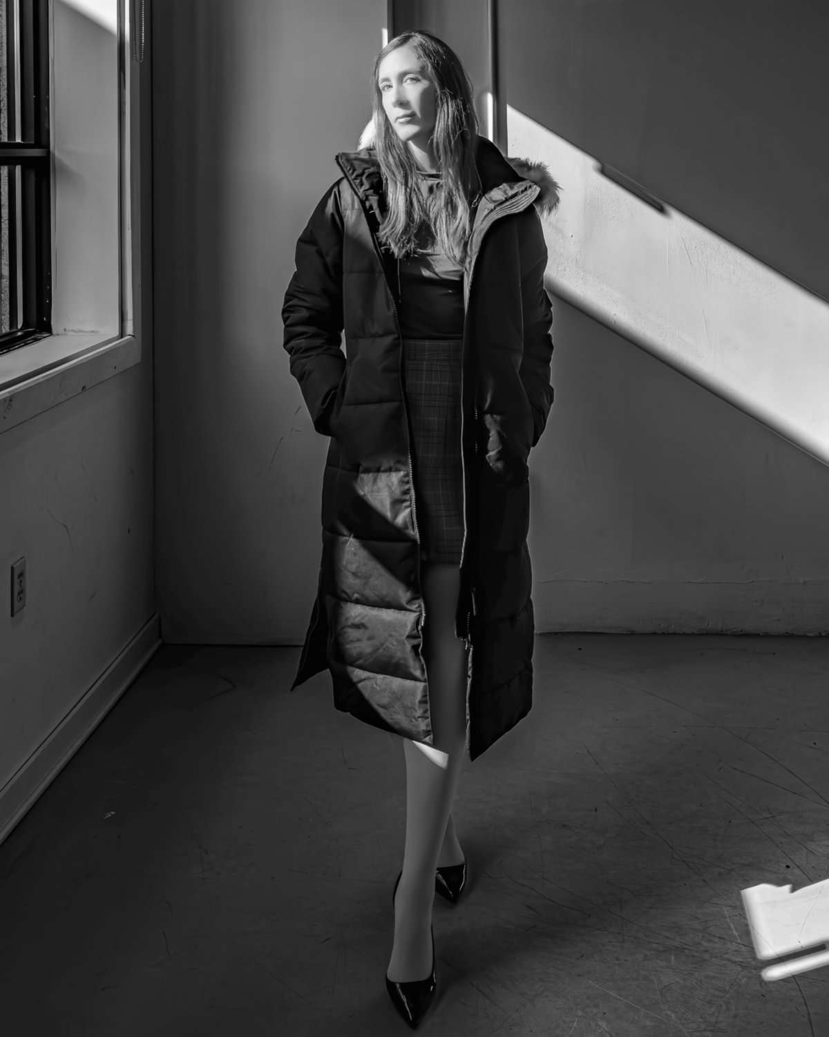 Laura Warner models a long black NOIZE puffer coat, paired with a plaid skirt, white tights, and black high heels, captured in a dramatic black and white fashion portrait with strong window light.