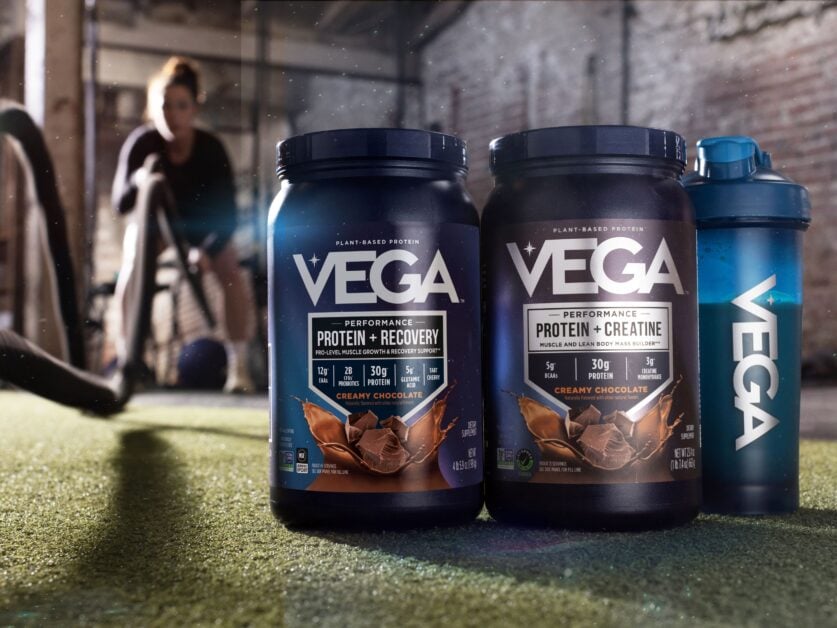 A side-by-side comparison of Vega Protein plus Recovery and Vega Protein plus Creatine tubs with a Vega shaker bottle in a gym setting.